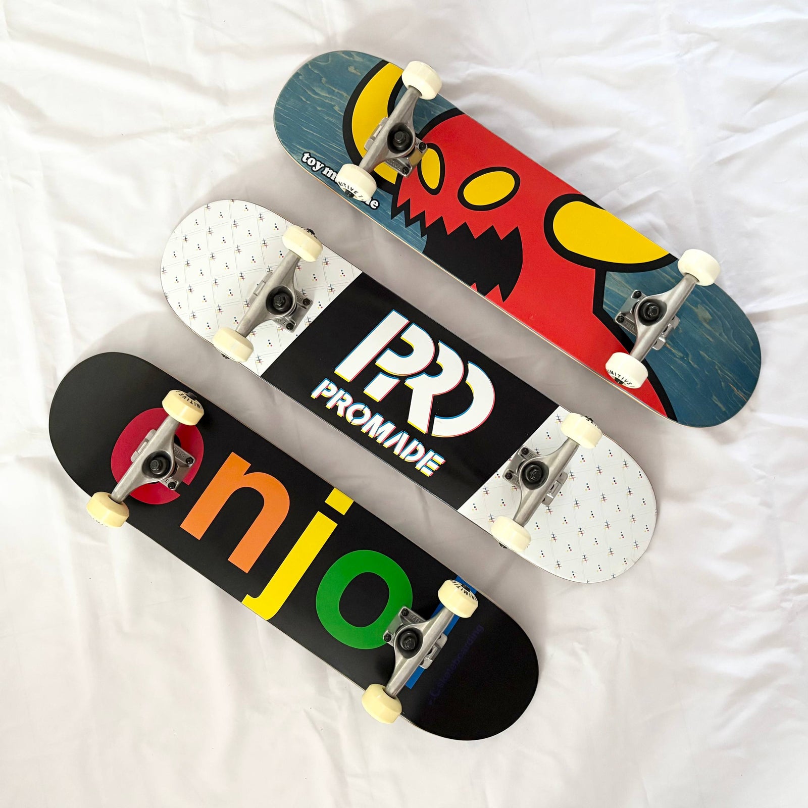 Three pre-built/custom complete skateboards from Toy Machine, Promade and Enjoi arranged diagonally on white fabric.