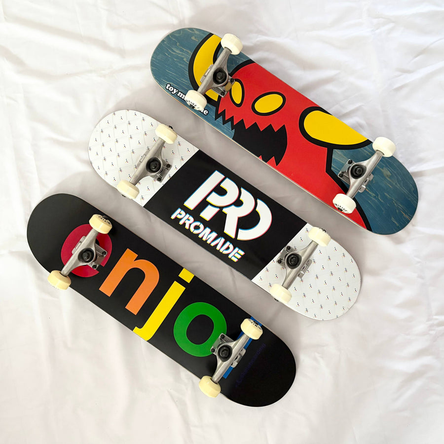 Three pre-built/custom complete skateboards from Toy Machine, Promade and Enjoi arranged diagonally on white fabric.